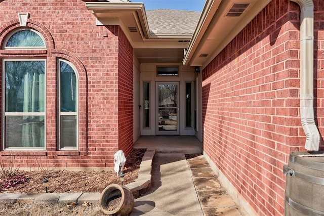 view of exterior entry featuring visible vents, brick siding, and roof with shingles