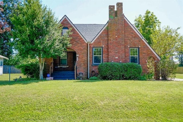 tudor-style house with brick siding, fence, roof with shingles, a front yard, and a chimney