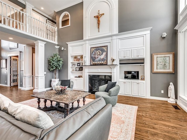 living area featuring wood finished floors, a fireplace, baseboards, a towering ceiling, and ornate columns