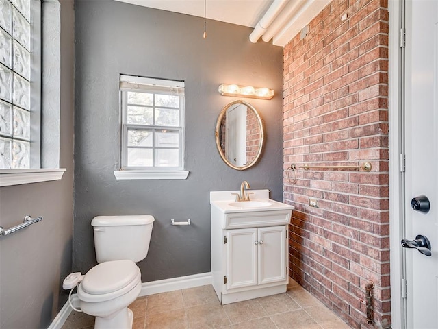 half bathroom featuring tile patterned flooring, brick wall, baseboards, toilet, and vanity