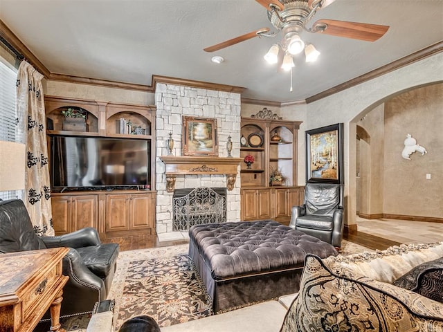 living room with a ceiling fan, wood finished floors, arched walkways, a stone fireplace, and crown molding