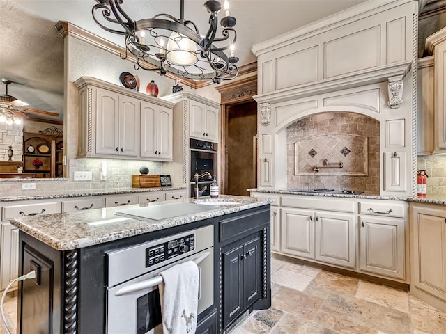 kitchen with tasteful backsplash, a center island with sink, ceiling fan with notable chandelier, stone tile flooring, and black electric cooktop