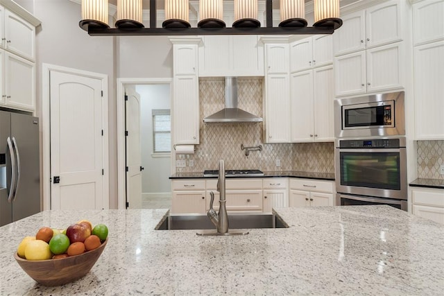 kitchen featuring a sink, light stone counters, tasteful backsplash, stainless steel appliances, and wall chimney range hood