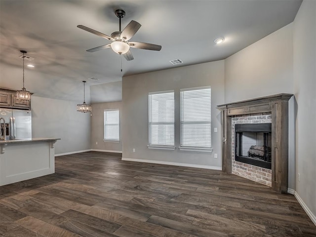 unfurnished living room with dark wood finished floors, a brick fireplace, ceiling fan with notable chandelier, and visible vents