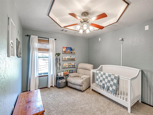 carpeted bedroom with a textured ceiling, a nursery area, a raised ceiling, ceiling fan, and a textured wall