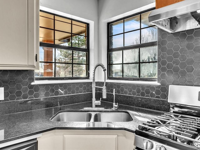 kitchen with backsplash, wall chimney range hood, gas range, white cabinets, and a sink