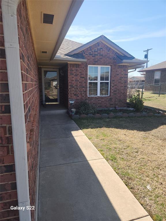 doorway to property with brick siding, a shingled roof, a lawn, and fence