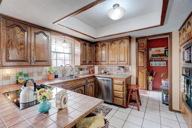 kitchen featuring tile countertops, a tray ceiling, decorative backsplash, stainless steel dishwasher, and a sink