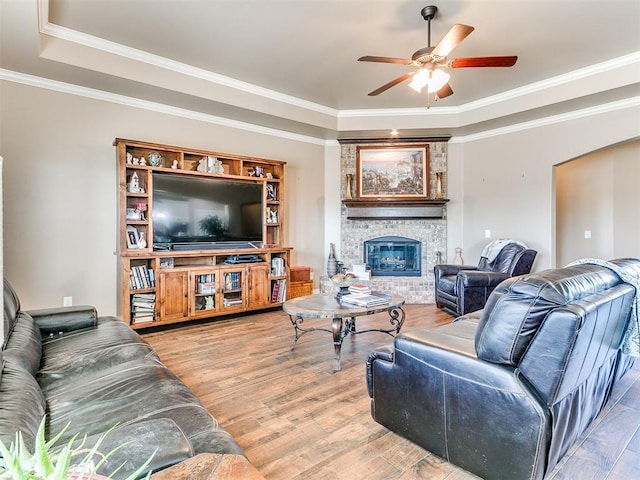 living room featuring light wood finished floors, a tray ceiling, ornamental molding, a fireplace, and a ceiling fan