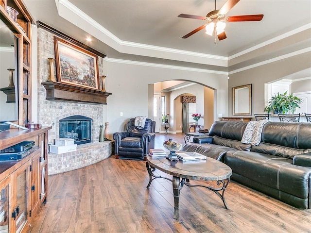 living area featuring a tray ceiling, wood finished floors, arched walkways, crown molding, and a brick fireplace