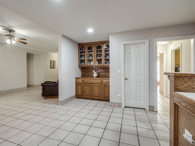 kitchen featuring light tile patterned floors, brown cabinetry, baseboards, ceiling fan, and glass insert cabinets