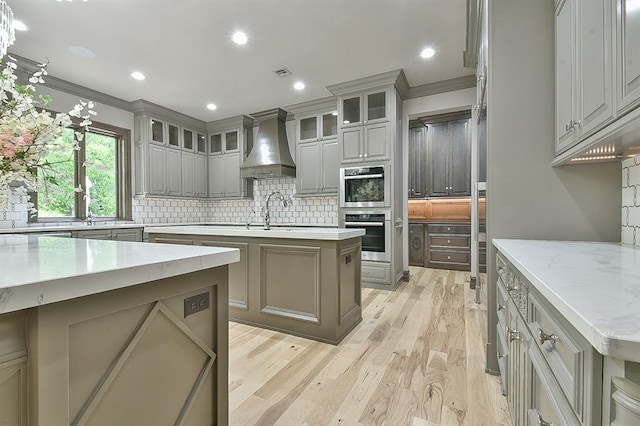 2747 Walking Woods Trl, Edmond, OK 73049 - kitchen with custom range hood, gray cabinets, decorative backsplash, light wood-style flooring, and a kitchen island with sink