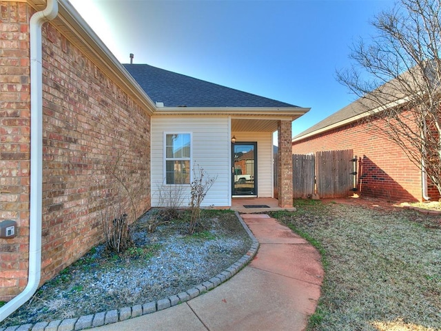 doorway to property with brick siding, a shingled roof, and fence