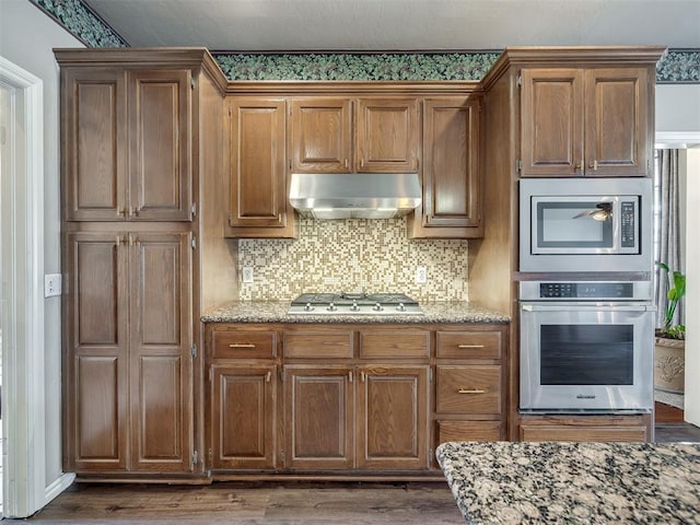 kitchen with light stone counters, dark wood-type flooring, under cabinet range hood, appliances with stainless steel finishes, and backsplash