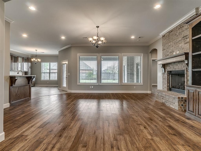 unfurnished living room featuring a notable chandelier, a brick fireplace, crown molding, and dark wood-style flooring