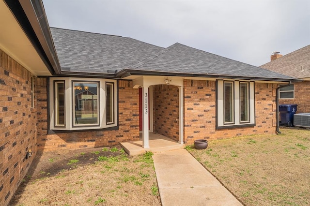 property entrance with central AC unit, brick siding, roof with shingles, and a lawn