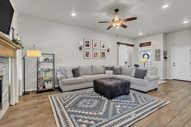 living area featuring ceiling fan, light wood-type flooring, a barn door, a stone fireplace, and recessed lighting