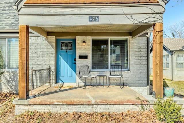 view of exterior entry with brick siding, covered porch, and a shingled roof