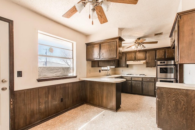 kitchen featuring stainless steel double oven, a peninsula, wainscoting, white gas stovetop, and under cabinet range hood