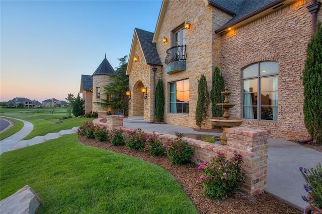 french country inspired facade featuring stone siding, a balcony, a shingled roof, and a front yard