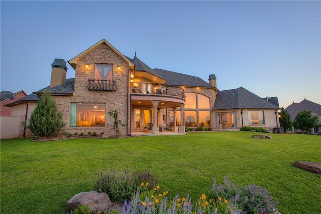 back of house at dusk with a lawn, a patio, a chimney, and a balcony