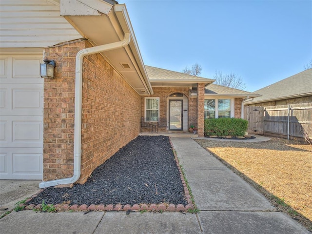 property entrance with brick siding, an attached garage, a shingled roof, and fence