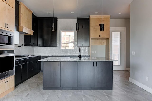 kitchen with light stone counters, an island with sink, a sink, decorative backsplash, and appliances with stainless steel finishes