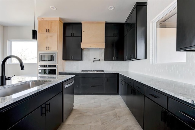 kitchen with dark cabinetry, light stone counters, a sink, custom range hood, and appliances with stainless steel finishes