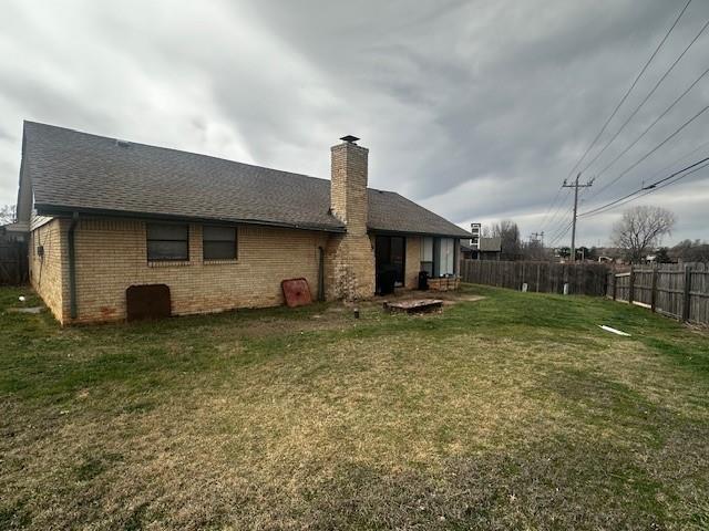 rear view of property with fence, a yard, roof with shingles, brick siding, and a chimney