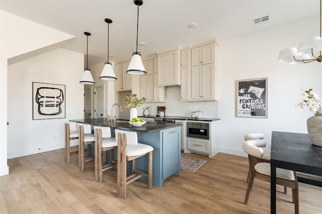 kitchen featuring visible vents, light wood-type flooring, a kitchen island with sink, backsplash, and dark countertops