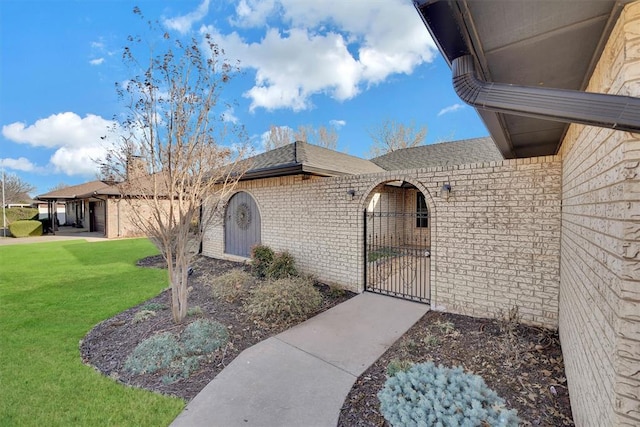 view of exterior entry featuring a gate, a lawn, brick siding, and a shingled roof