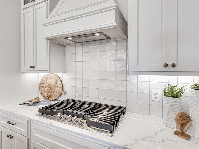 kitchen featuring light stone countertops, premium range hood, stainless steel gas cooktop, white cabinets, and backsplash