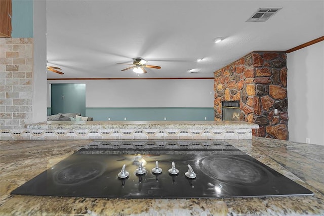 kitchen featuring visible vents, a ceiling fan, a stone fireplace, and crown molding