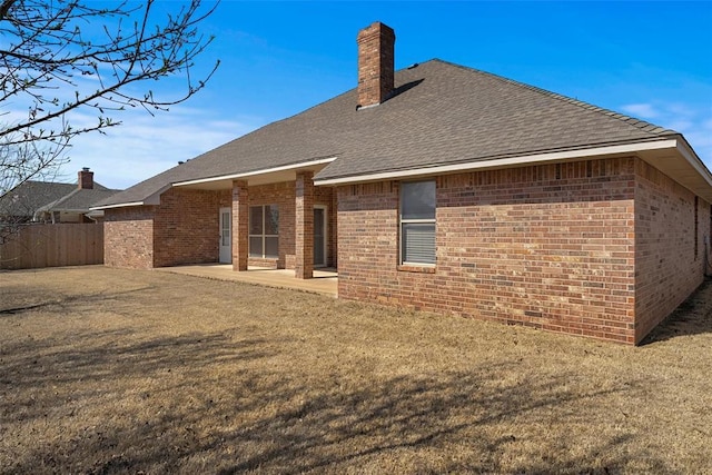 rear view of property featuring fence, roof with shingles, brick siding, a chimney, and a patio area