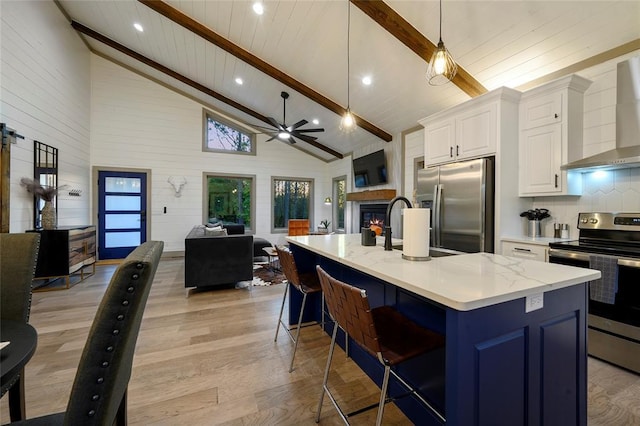 kitchen featuring beam ceiling, open floor plan, stainless steel appliances, wall chimney range hood, and ceiling fan