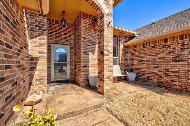 view of exterior entry with brick siding, roof with shingles, and a patio area