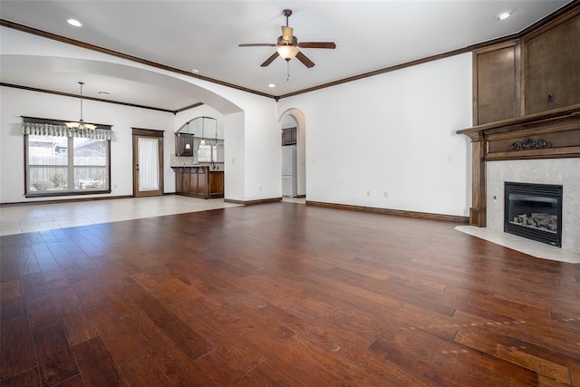 unfurnished living room with baseboards, arched walkways, hardwood / wood-style flooring, a tiled fireplace, and ceiling fan with notable chandelier