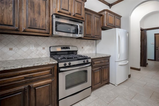 kitchen featuring light stone countertops, dark brown cabinetry, decorative backsplash, appliances with stainless steel finishes, and arched walkways