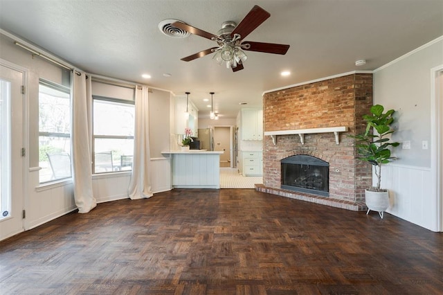 unfurnished living room featuring a ceiling fan, a fireplace, wainscoting, crown molding, and a decorative wall