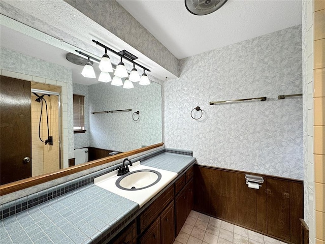 bathroom with a wainscoted wall, a textured ceiling, a tile shower, wallpapered walls, and vanity