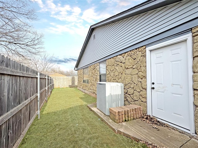 view of exterior entry featuring stone siding, a lawn, central AC, and fence