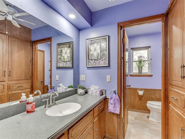 bathroom with wood walls, toilet, wainscoting, vanity, and a ceiling fan