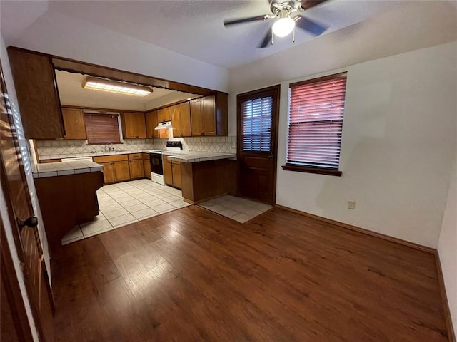 kitchen featuring light wood finished floors, backsplash, tile countertops, brown cabinetry, and white electric range