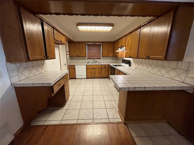 kitchen with electric range, white dishwasher, tile counters, under cabinet range hood, and brown cabinets