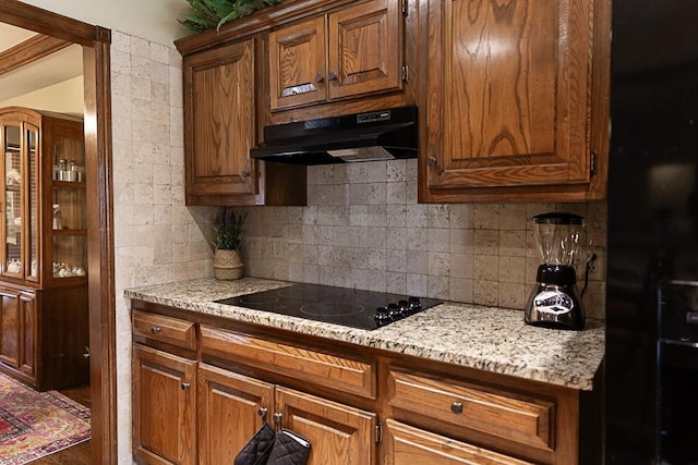 kitchen featuring light stone counters, brown cabinets, under cabinet range hood, black electric cooktop, and tasteful backsplash