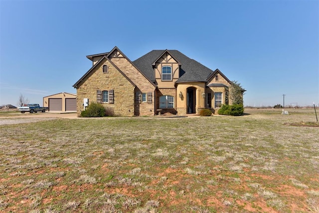 french country inspired facade with a garage, stone siding, an outdoor structure, and a front yard