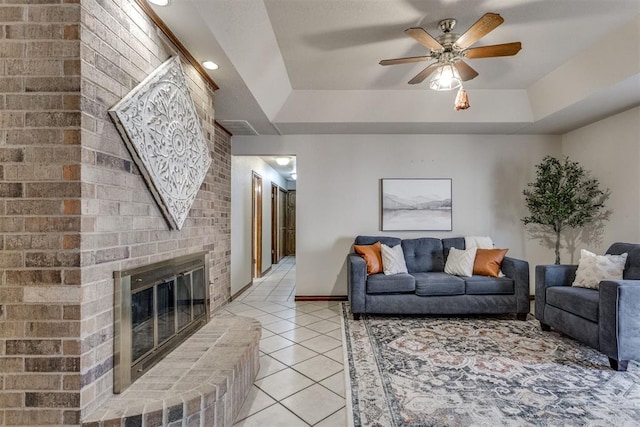 living area with a tray ceiling, a brick fireplace, light tile patterned flooring, and ceiling fan