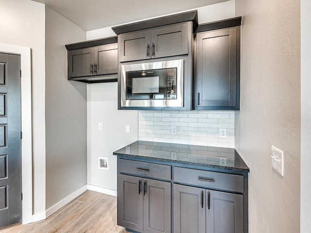 kitchen featuring baseboards, light wood finished floors, dark stone counters, decorative backsplash, and stainless steel microwave