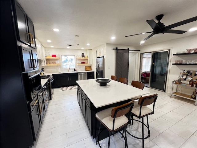 kitchen with dark cabinetry, a breakfast bar area, open shelves, black appliances, and a barn door