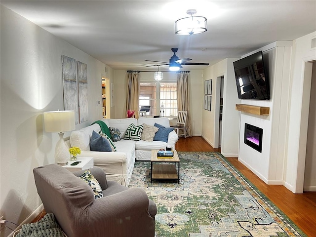 living room featuring a glass covered fireplace, wood finished floors, baseboards, and ceiling fan